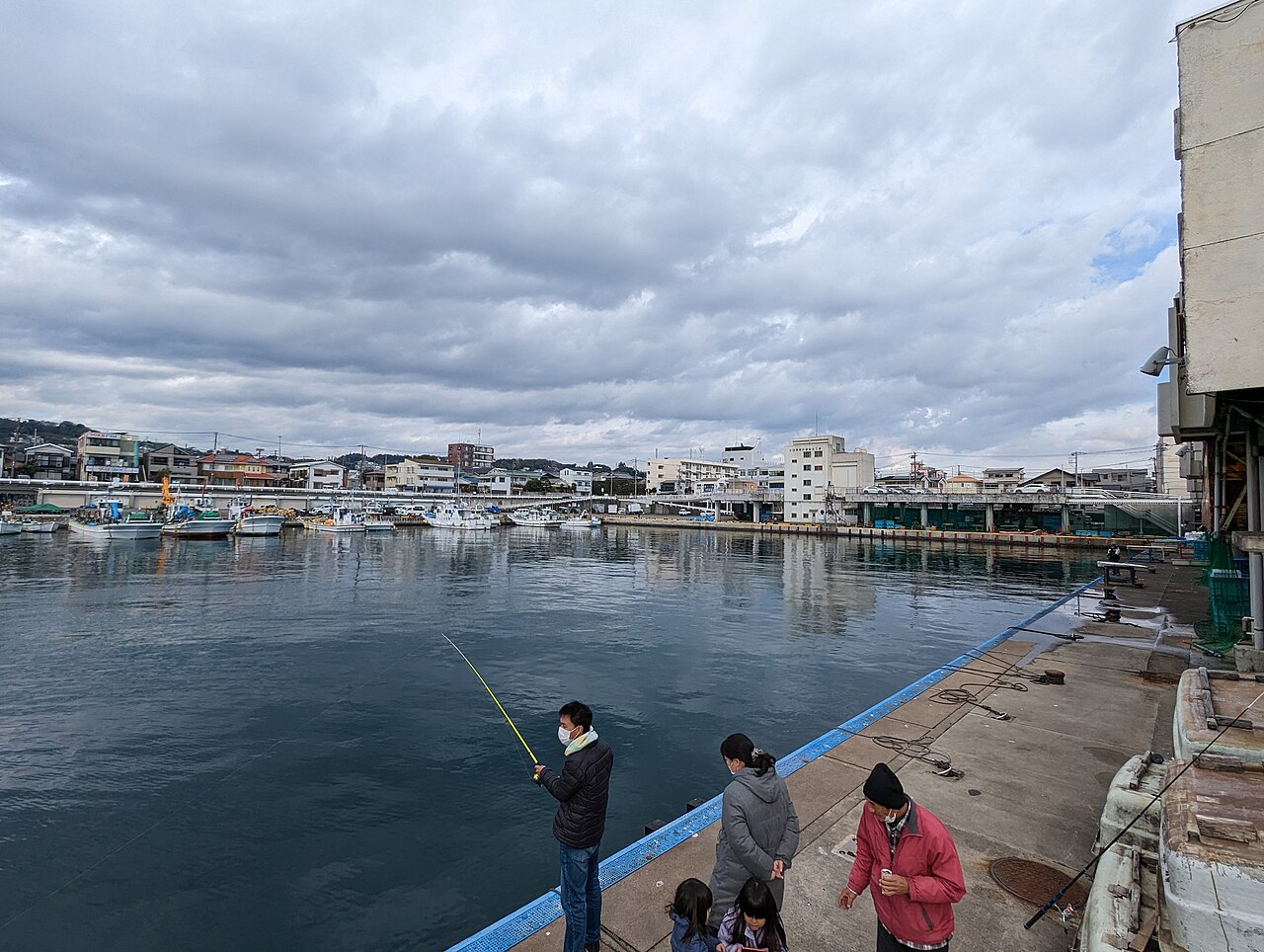 The calm water of Odawara fishing port with small fishing boats moored along the far quay under a cloudy sky, and a few people fishing from the near quay