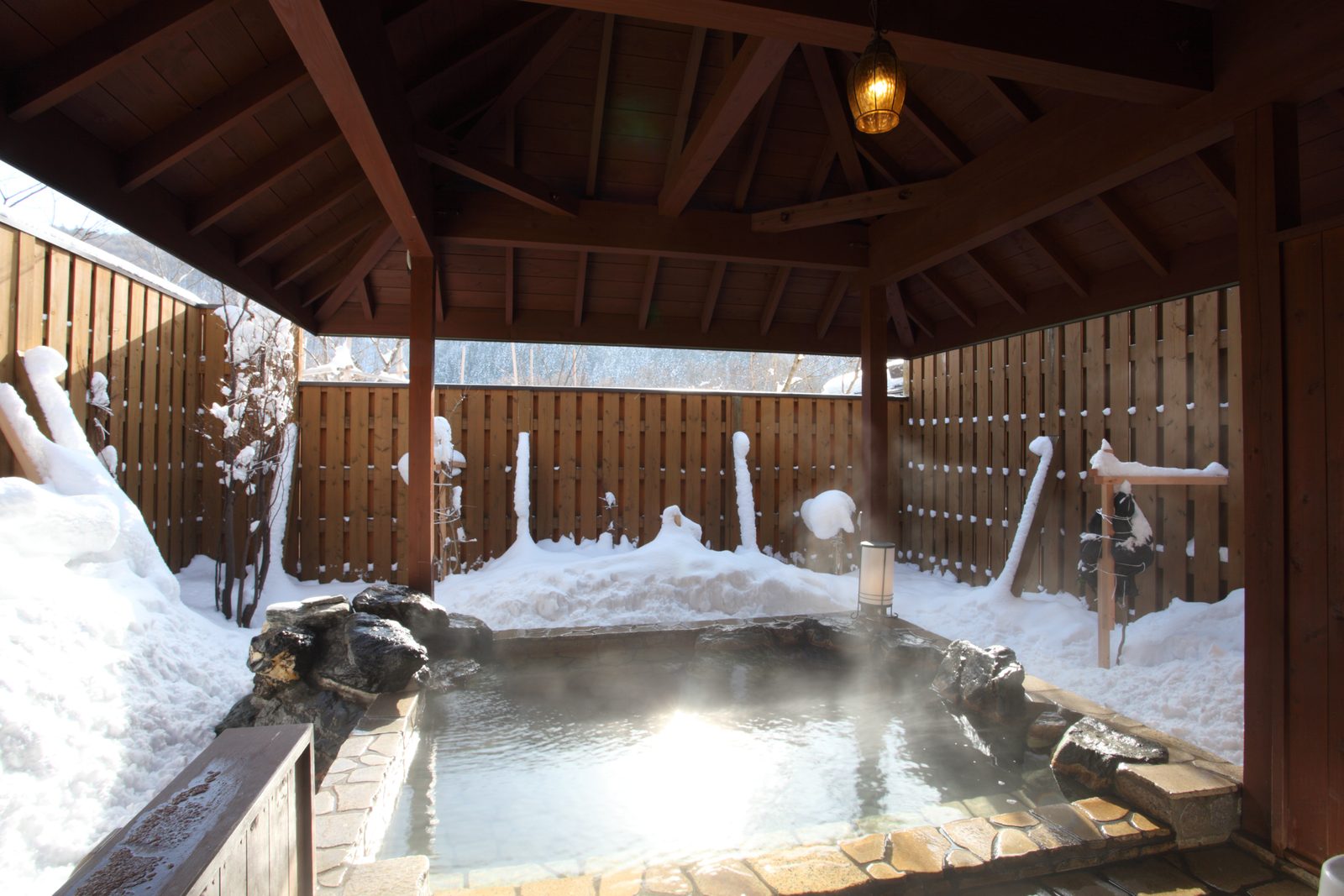 A snow-ringed outdoor hot spring bath under a wooden pavilion roof near Lake Towada, with steam rising off the water and stacked stones at the edge.