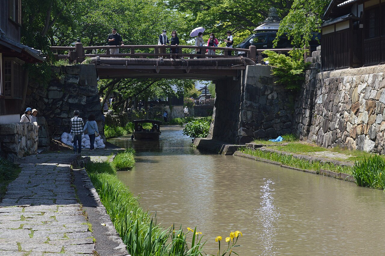 A stone-walled canal in Omihachiman with a wooden pleasure boat gliding toward an arched wooden bridge, old wooden townhouses and green trees along the banks.