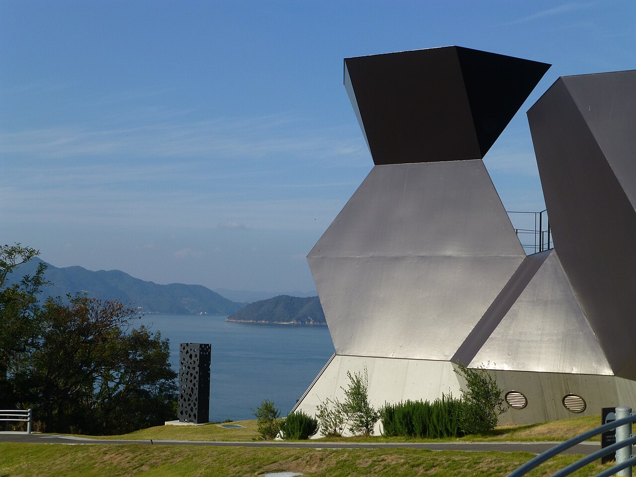 A faceted silver steel structure with a dark top stands on a green hill overlooking a blue sea and wooded islands