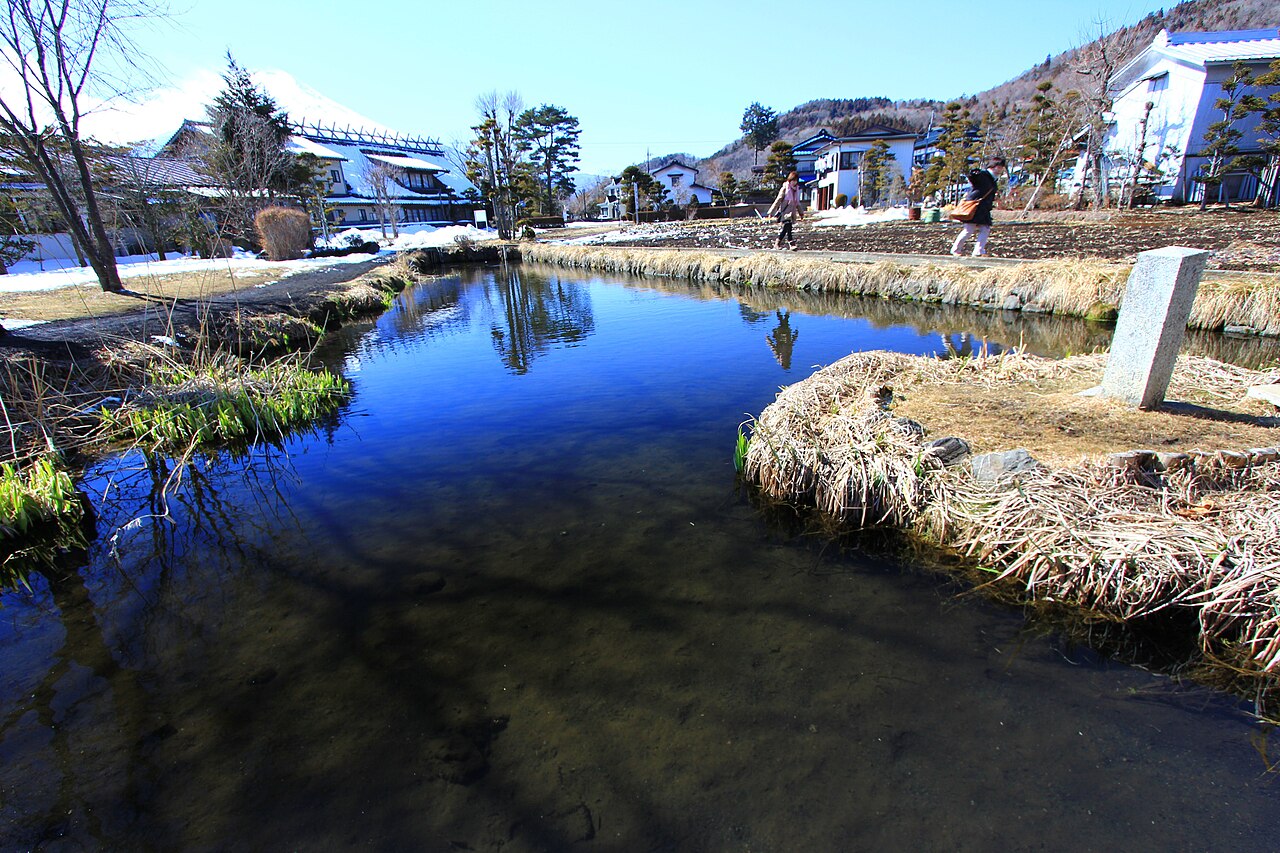 A clear spring-fed pond at Oshino Hakkai on a winter day, with village houses and mountains reflected in the still water beyond a stone marker