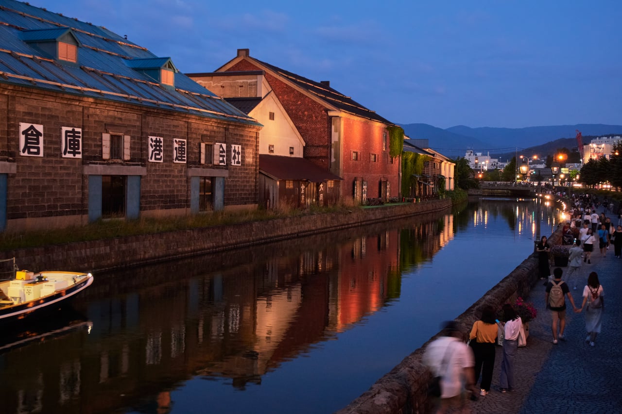 The Otaru Canal at dusk, lined with historic stone warehouses and gas lamps, with people strolling along the towpath.