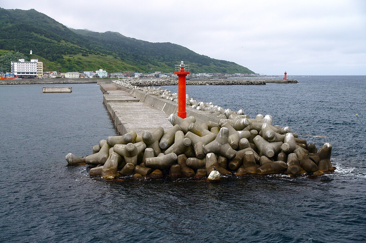 A concrete breakwater with a small red lighthouse and wave-breaker blocks at Kafuka Port, with green hills and the harbor town beyond.
