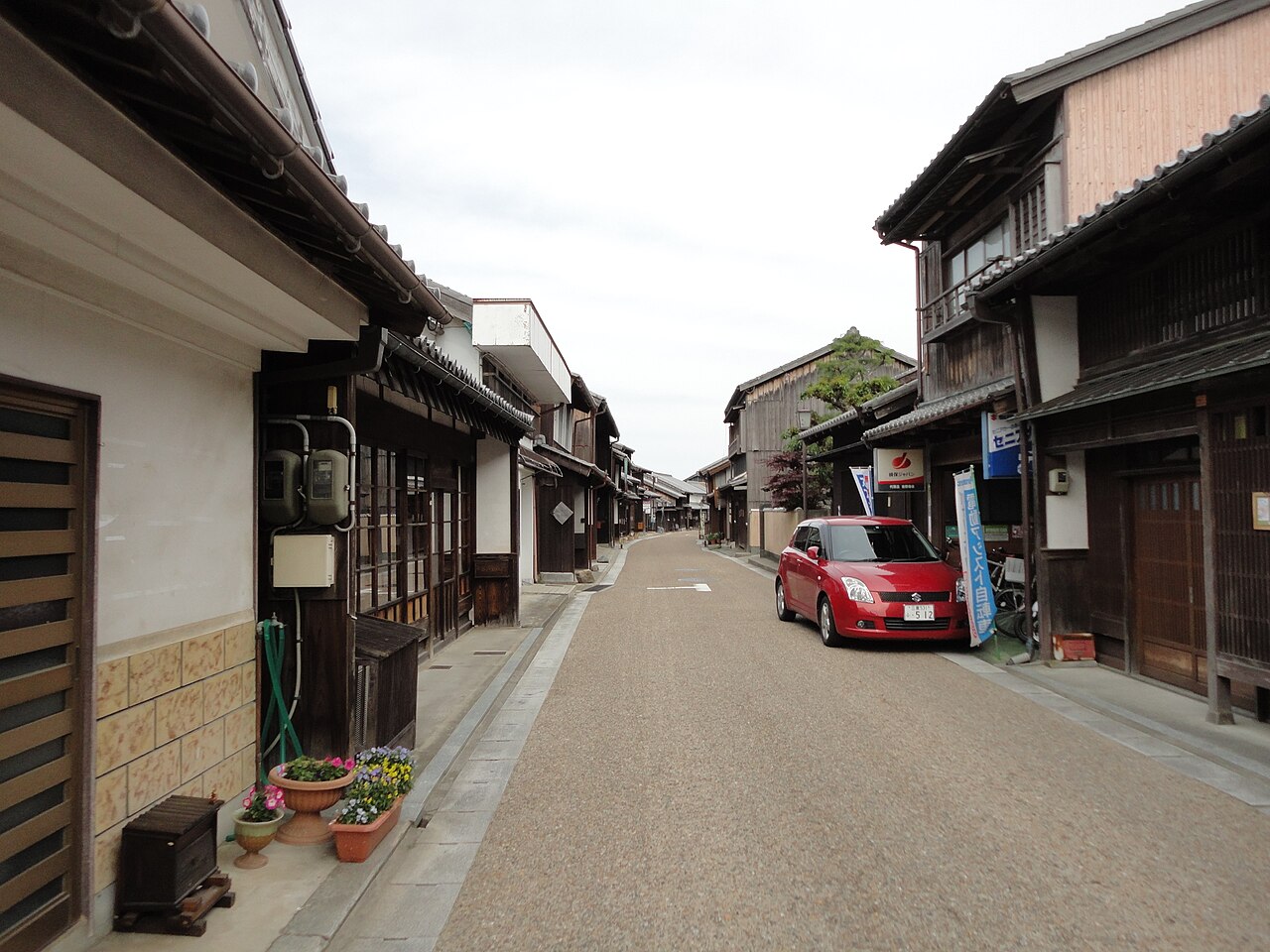 A narrow street of old wooden houses with tiled roofs and latticed fronts in Seki-juku, with a red car parked on the right.