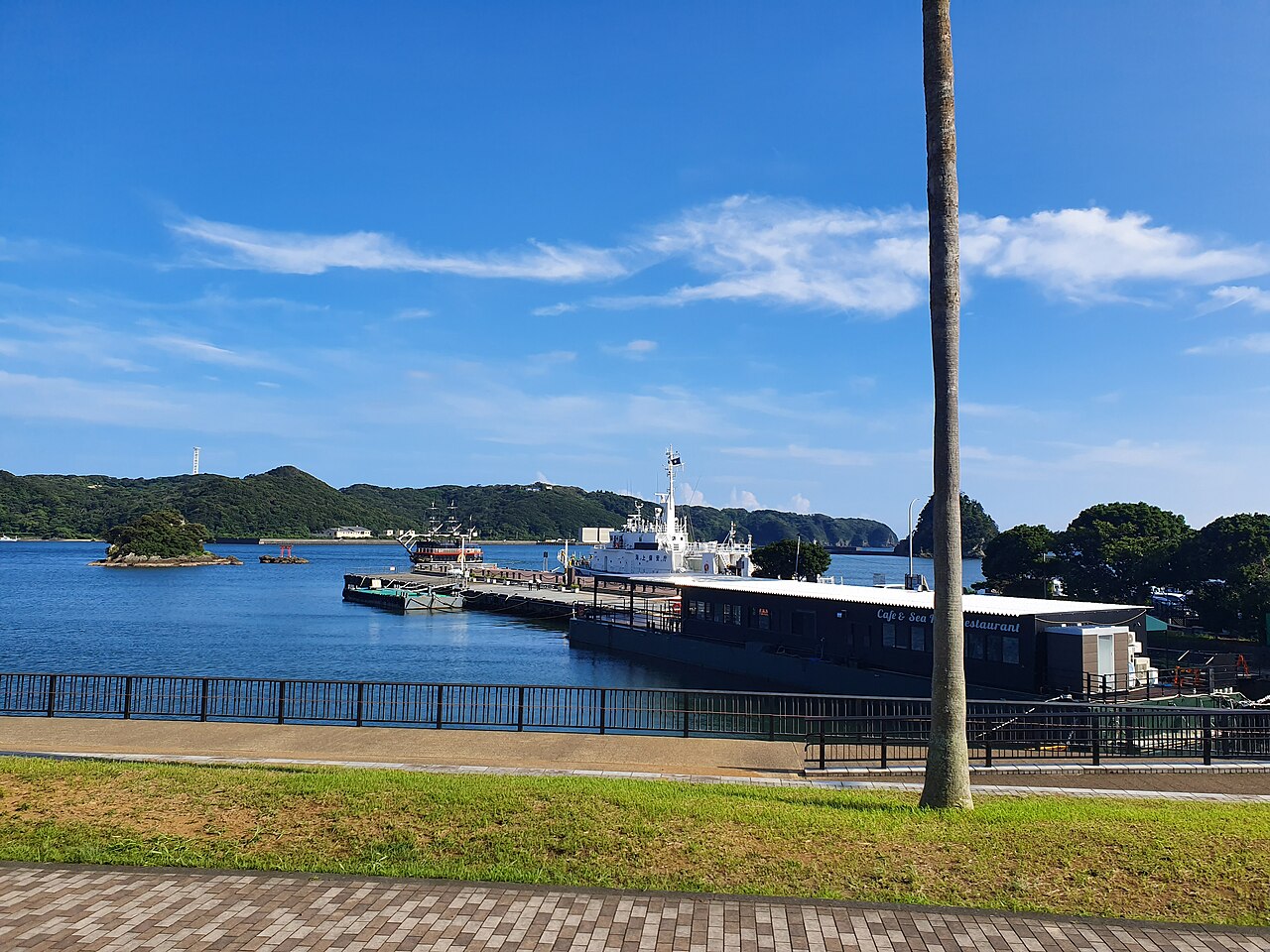 A calm harbor under a blue sky with a ferry-type boat moored at a pier, a lawn and brick-paved walkway in front, and small green islets across the water.