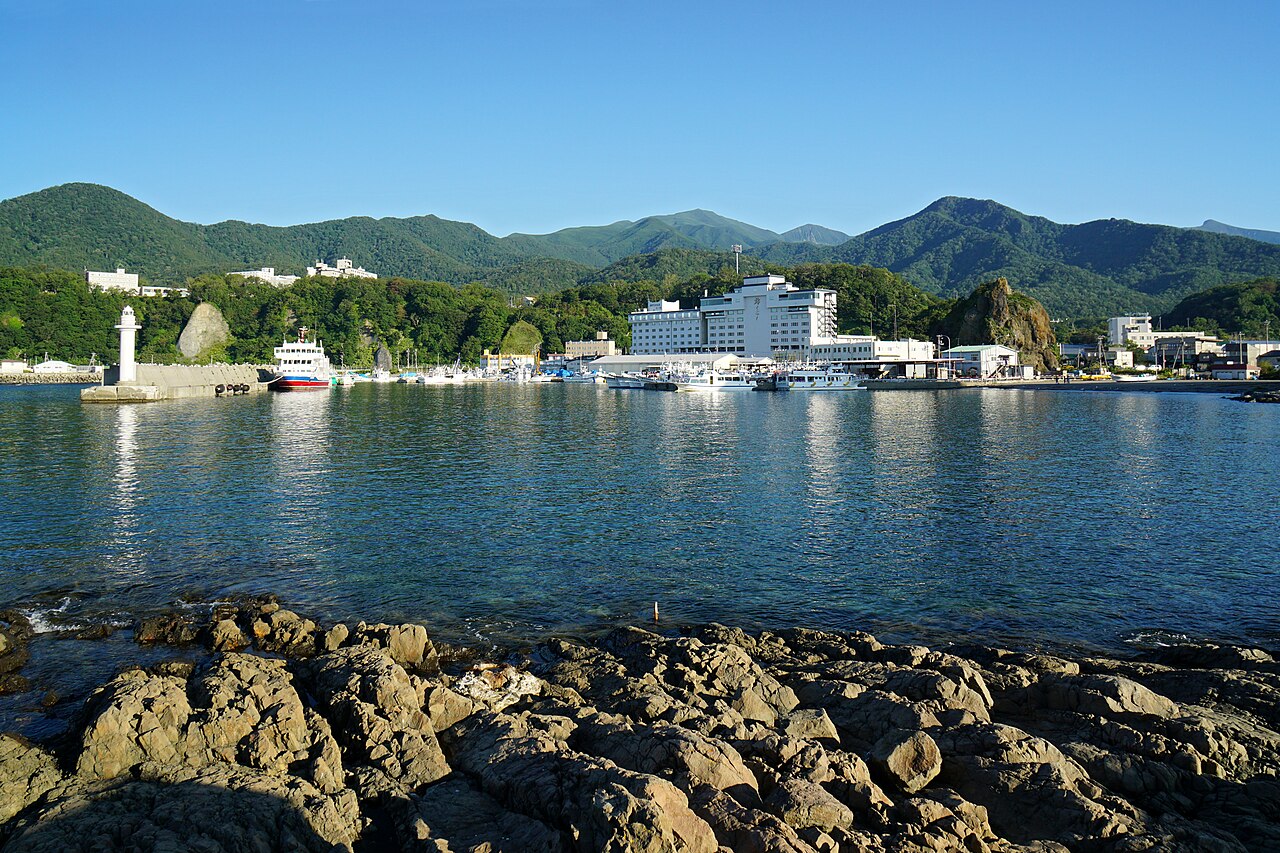 The small fishing harbor of Utoro on a calm bay with green mountains and a few low buildings along the shore.