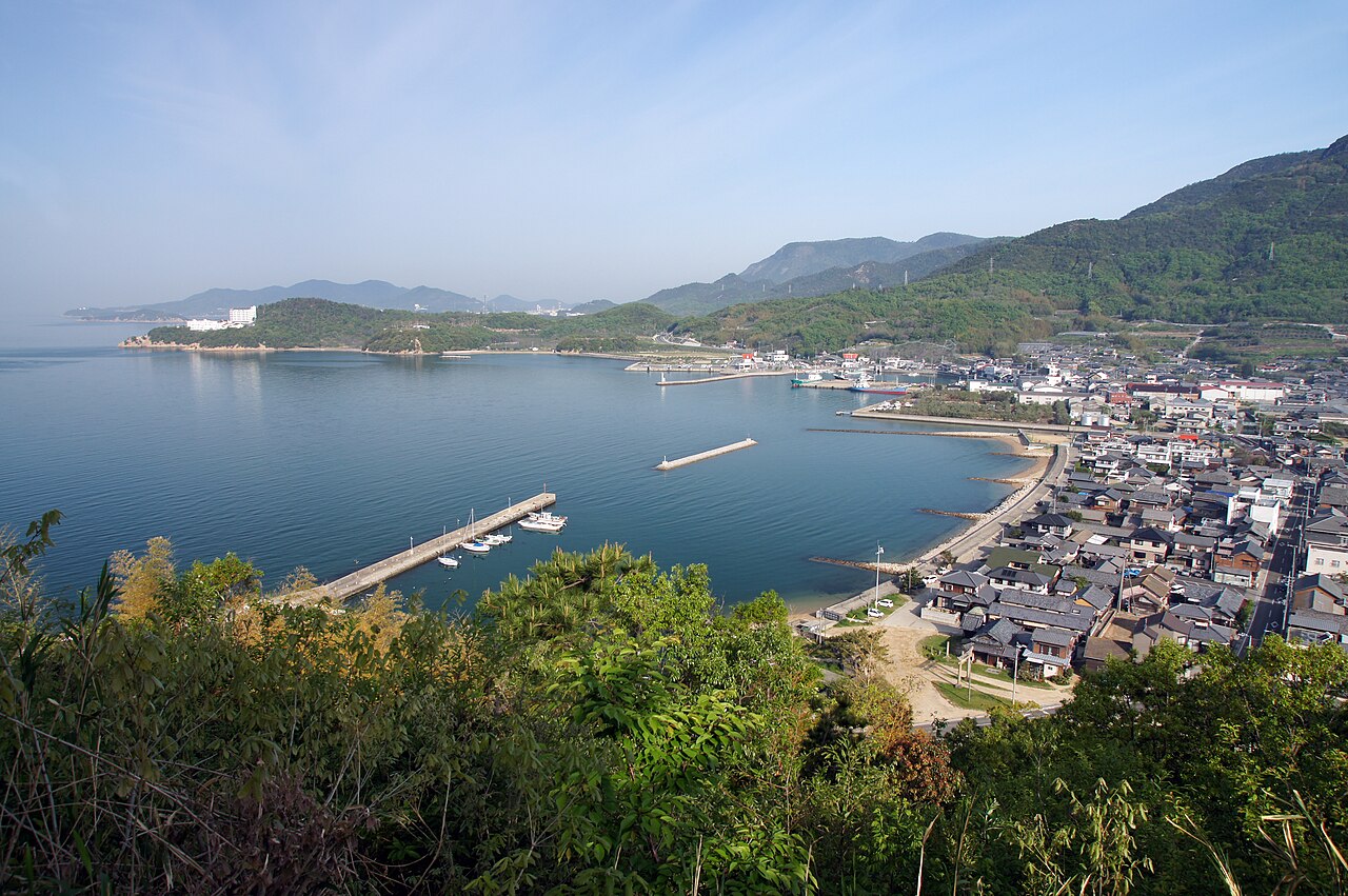 A quiet harbour on Shodoshima seen from a hillside, with a small jetty, moored boats, tiled-roof houses along the shore and green hills beyond