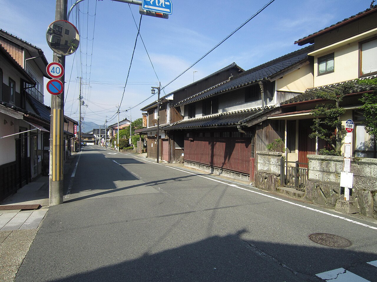 A quiet street of traditional dark-tiled wooden townhouses with a bus stop sign on the right, under a blue sky.