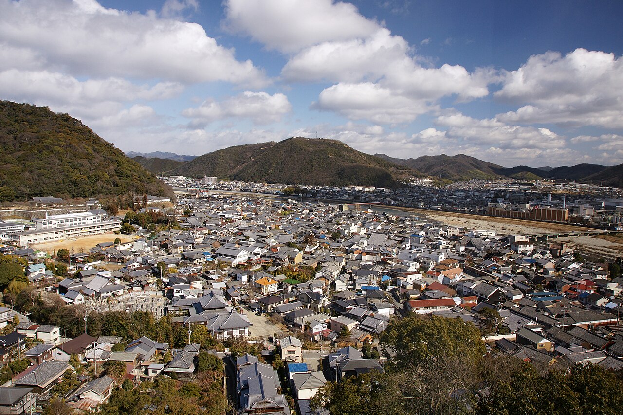 The roofs of the castle town of Tatsuno, Hyogo, seen from a hillside, with a river and wooded mountains beyond under a cloudy blue sky.