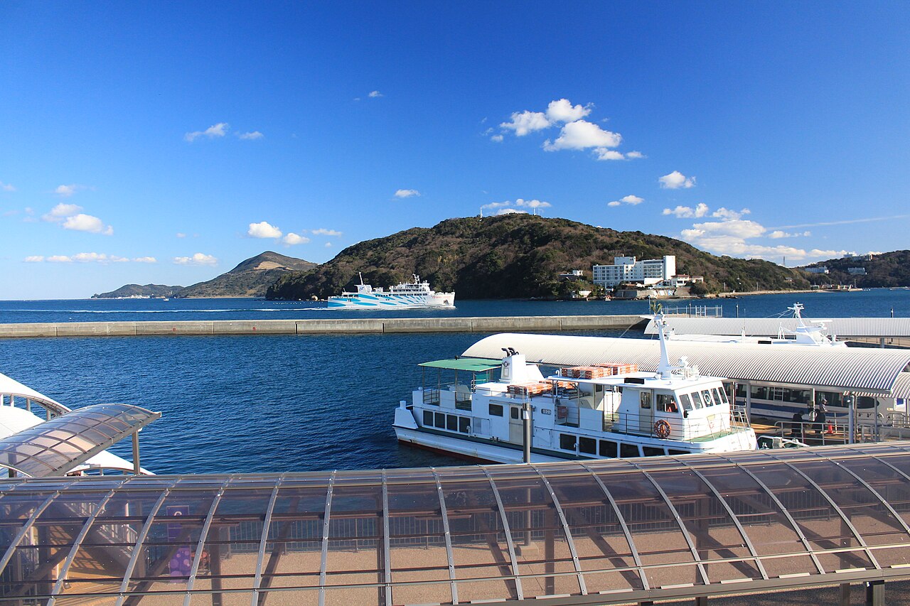 A green island and ferries moored at the Toba waterfront under a blue sky with hills across the bay