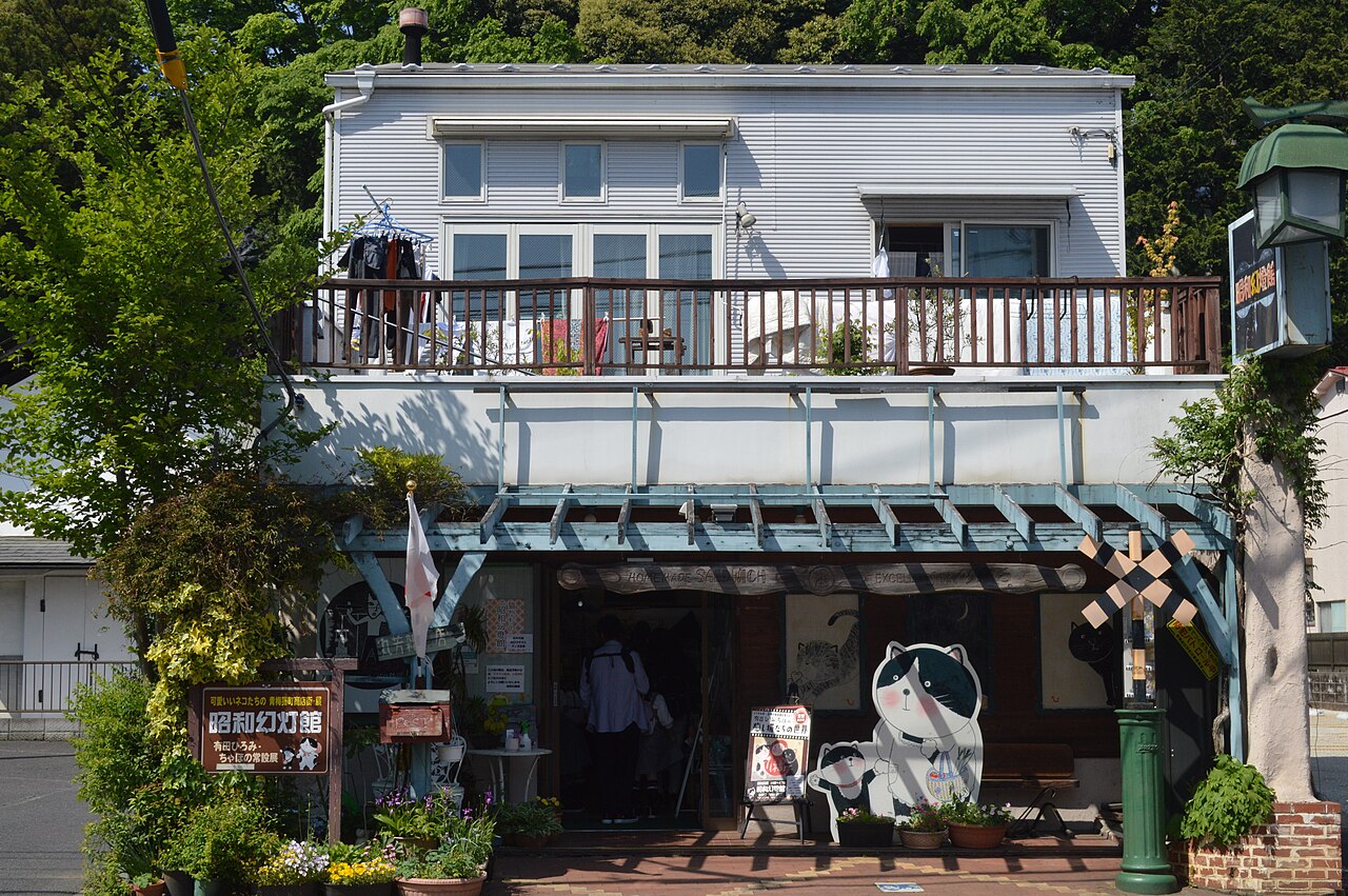 A small shop front with a blue wooden trellis canopy, flower pots, a cat cut-out and a miniature railway-crossing sign, below a balcony with a tree-covered hillside behind.