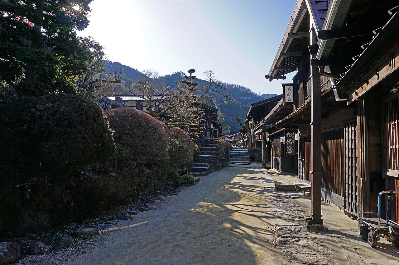 A sunlit gravel lane in Tsumago-juku between a stone wall with steps and a row of old wooden buildings, with hills in the distance