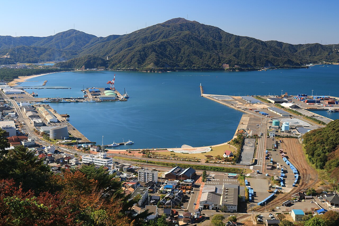 An aerial view of Tsuruga harbor with calm blue water, moored boats, and green mountains rising behind the waterfront town.