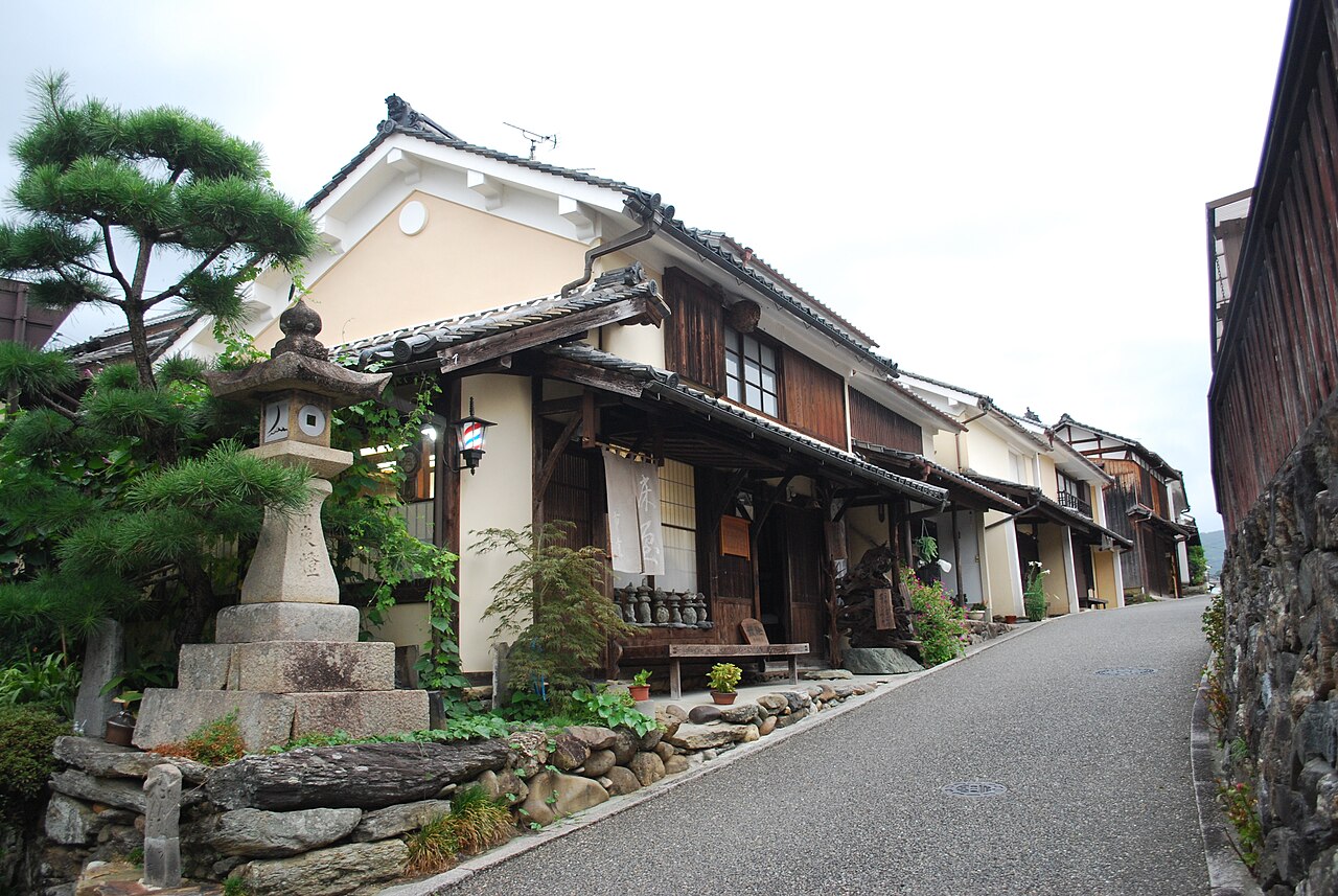 A sloping paved lane lined with old wooden and plaster townhouses in Uchiko, with a stone lantern and pine tree at the left