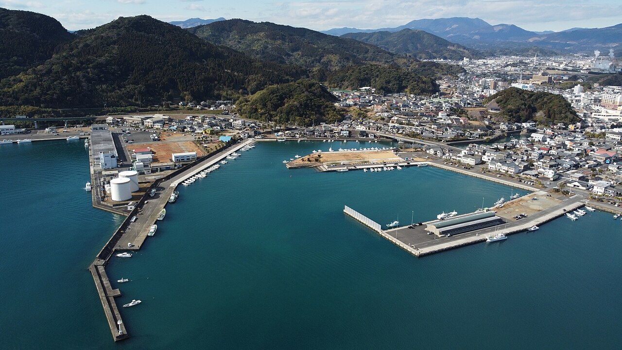 An aerial view of Aburatsu harbor and the surrounding town, with calm teal water, breakwaters, small boats and forested hills behind.