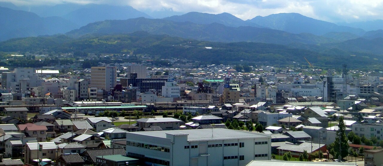 A town of low rooftops and mid-rise buildings spreading below a long forested mountain range under a broad sky.