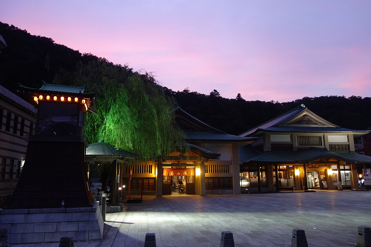 The Kikunoyu public bathhouse and a lantern tower in the Yamanaka Onsen town square at dusk, with a lit willow tree and forested hills behind.