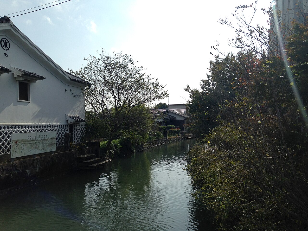 A quiet Yanagawa canal beside a white-walled storehouse with lattice-pattern lower walls, trees overhanging the water