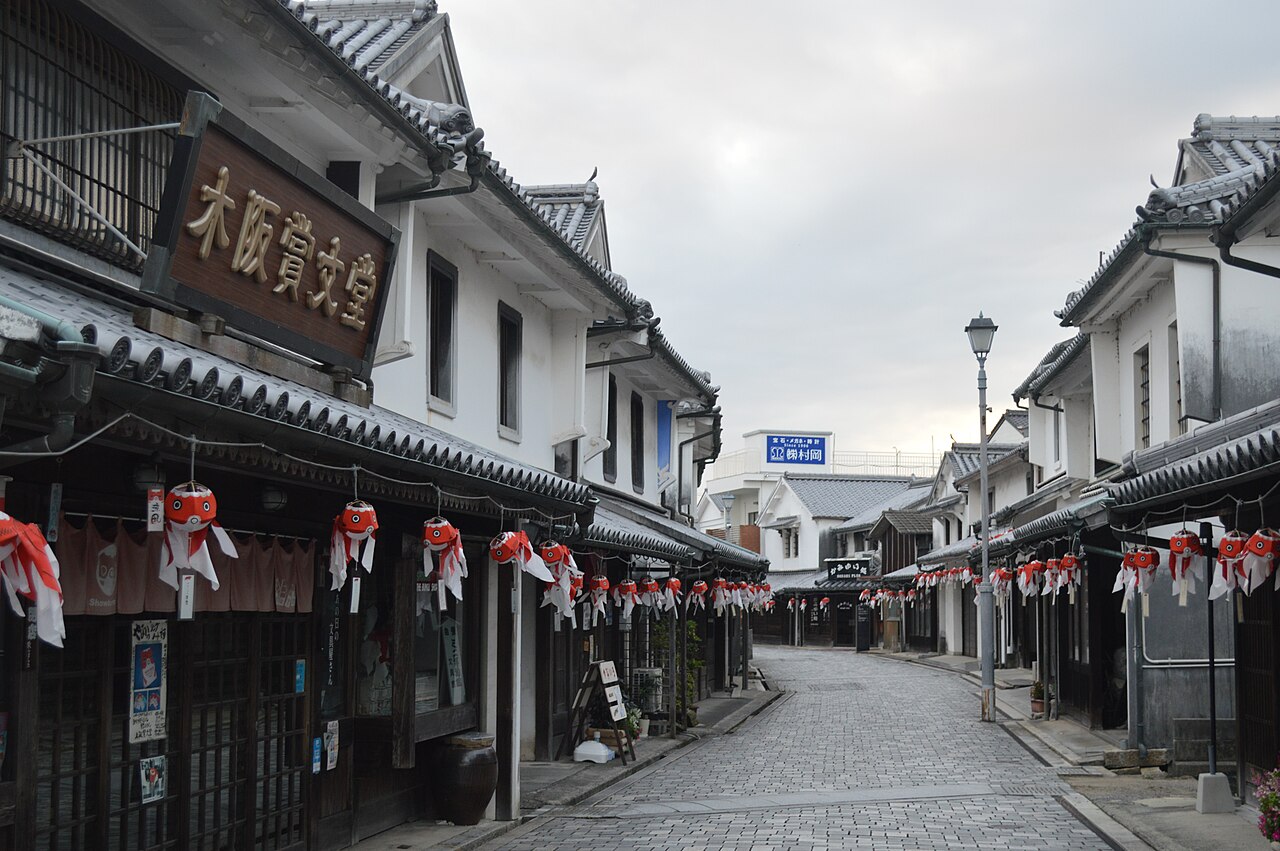 Quiet cobbled lane between white-walled merchant houses in the Yanai white-wall district under an overcast evening sky, with red goldfish lanterns hanging under the eaves
