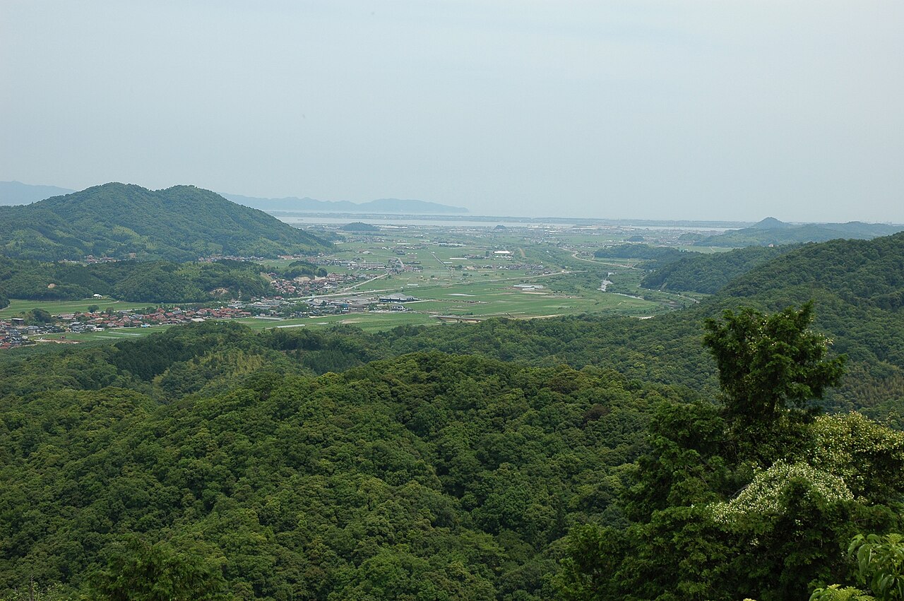 Wooded hills above a green plain with villages and a hazy lake beyond, seen from a hilltop