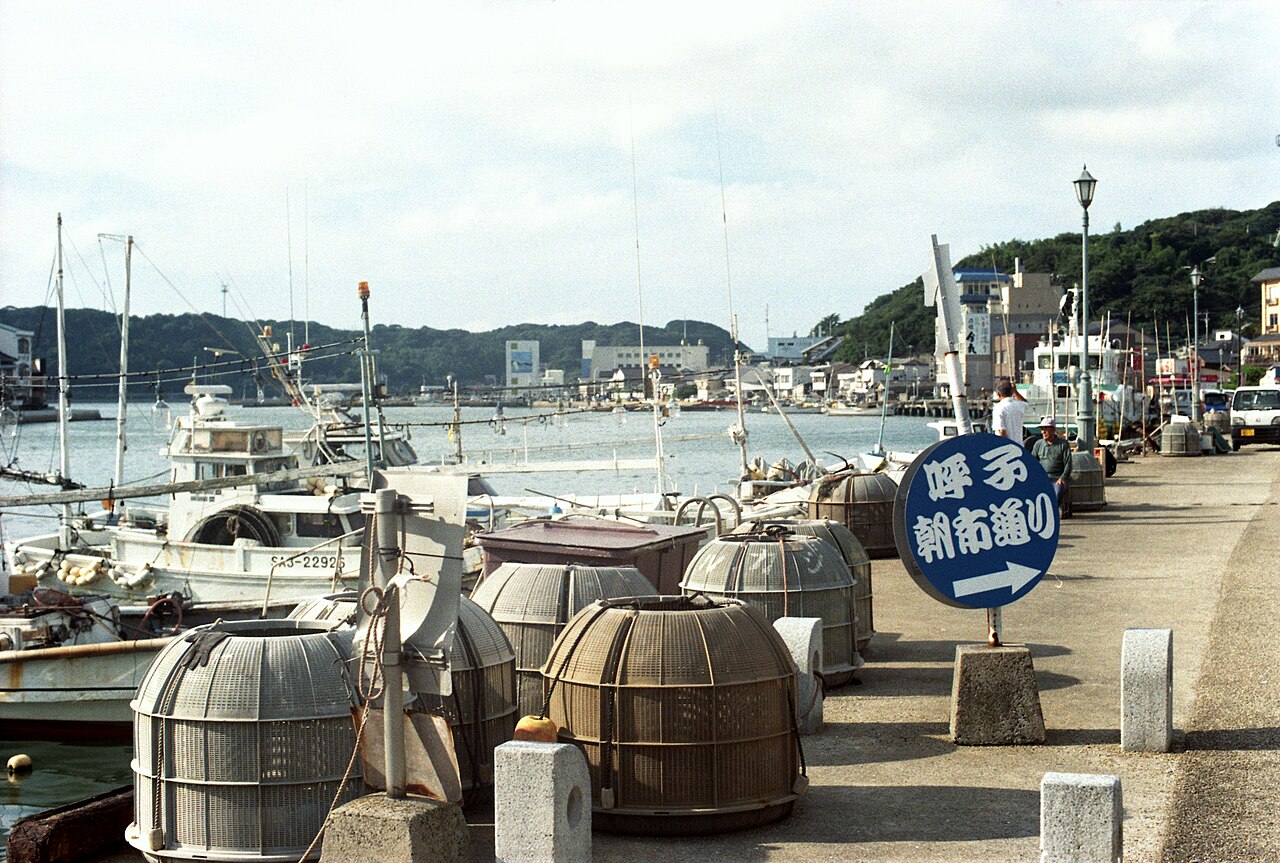 Fishing boats moored along a quay in Yobuko Port, with round baskets on the pavement and a blue sign pointing toward the morning market street