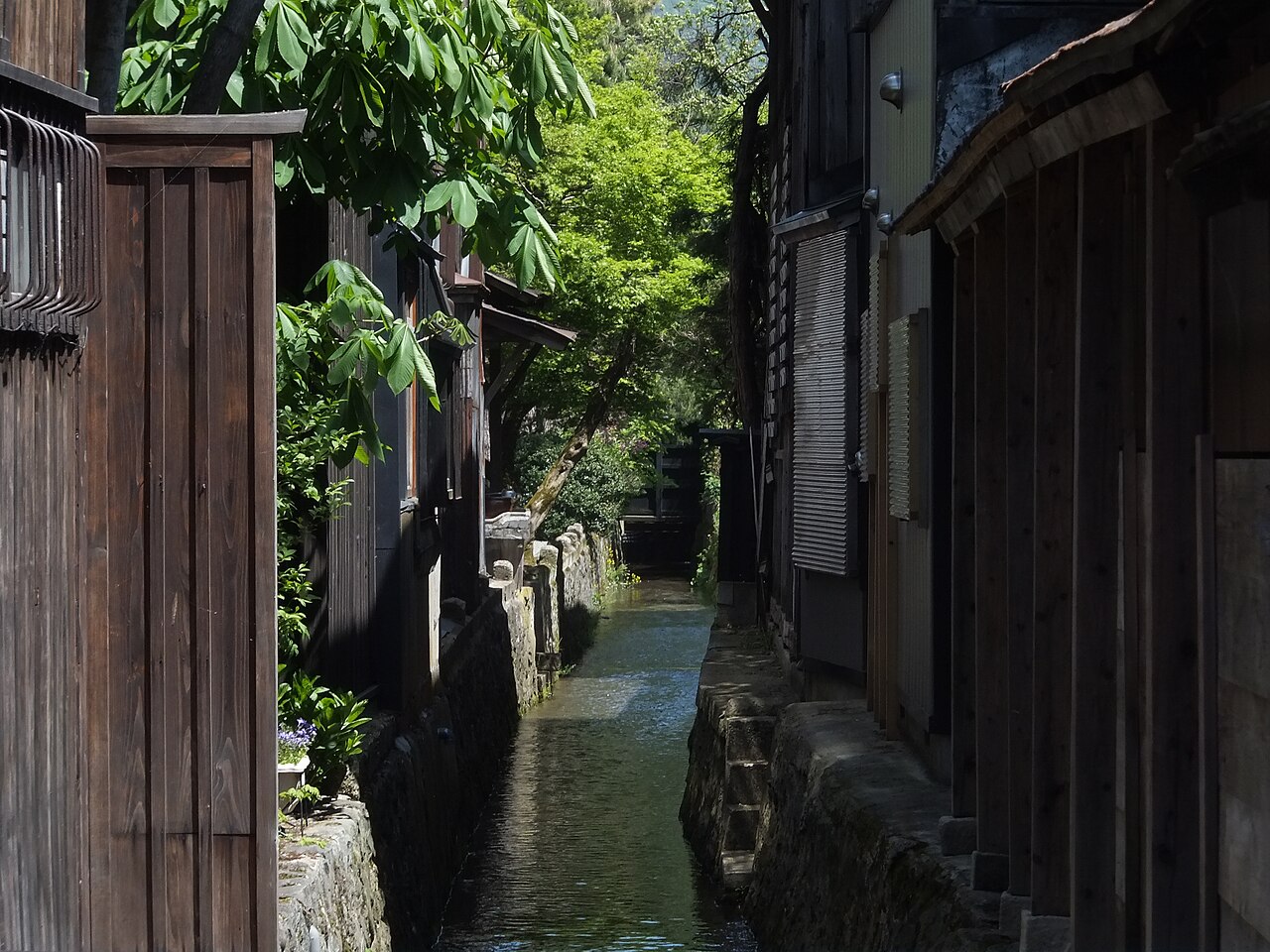 A narrow stone-lined water channel running between old wooden buildings and fences in a quiet town.