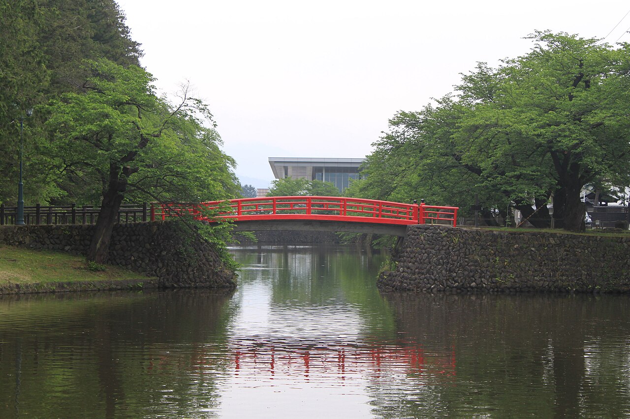 A red-railed arched bridge over the stone-walled moat beside Uesugi Shrine in Yonezawa, with leafy trees on both banks and a modern building behind.