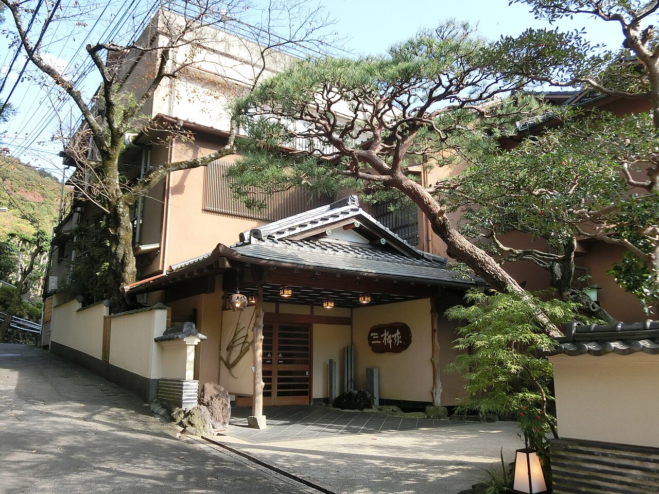 The wooden entrance of a traditional inn, with a tiled canopy, lanterns and a large pine tree leaning over a quiet paved lane.