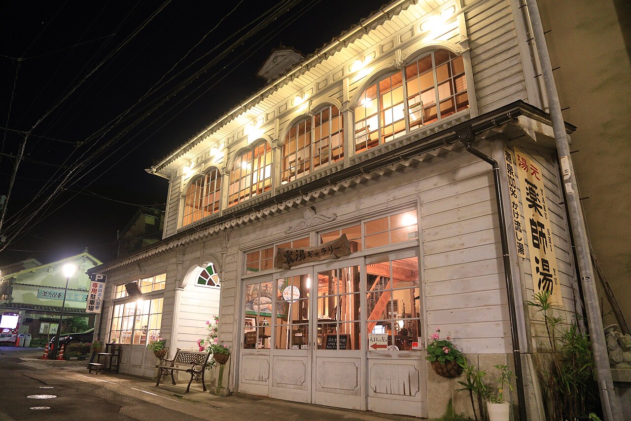 A Taisho-era wooden bathhouse building lit up at night on a quiet onsen street, with arched windows glowing warmly.