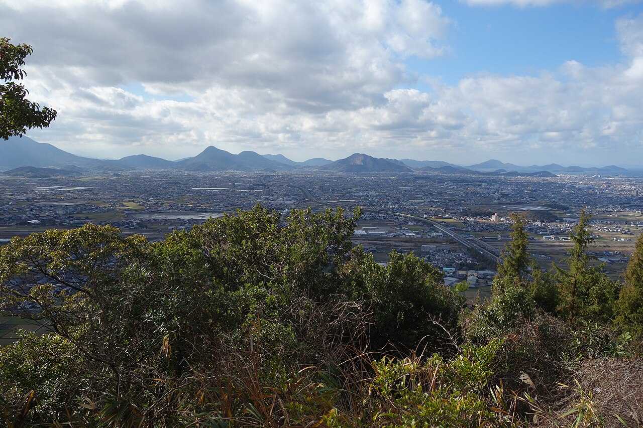 A wide view from a hilltop over the Sanuki plain, with towns and fields spreading to a row of low mountains under a bright, cloudy sky.