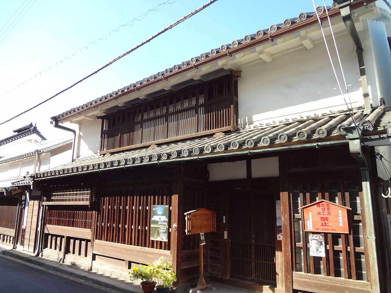 The Takagi House in Imai-cho, a two-storey wooden merchant house with white plaster walls, dark timber lattice fronts, and a red no-smoking notice board mounted beside the entrance.