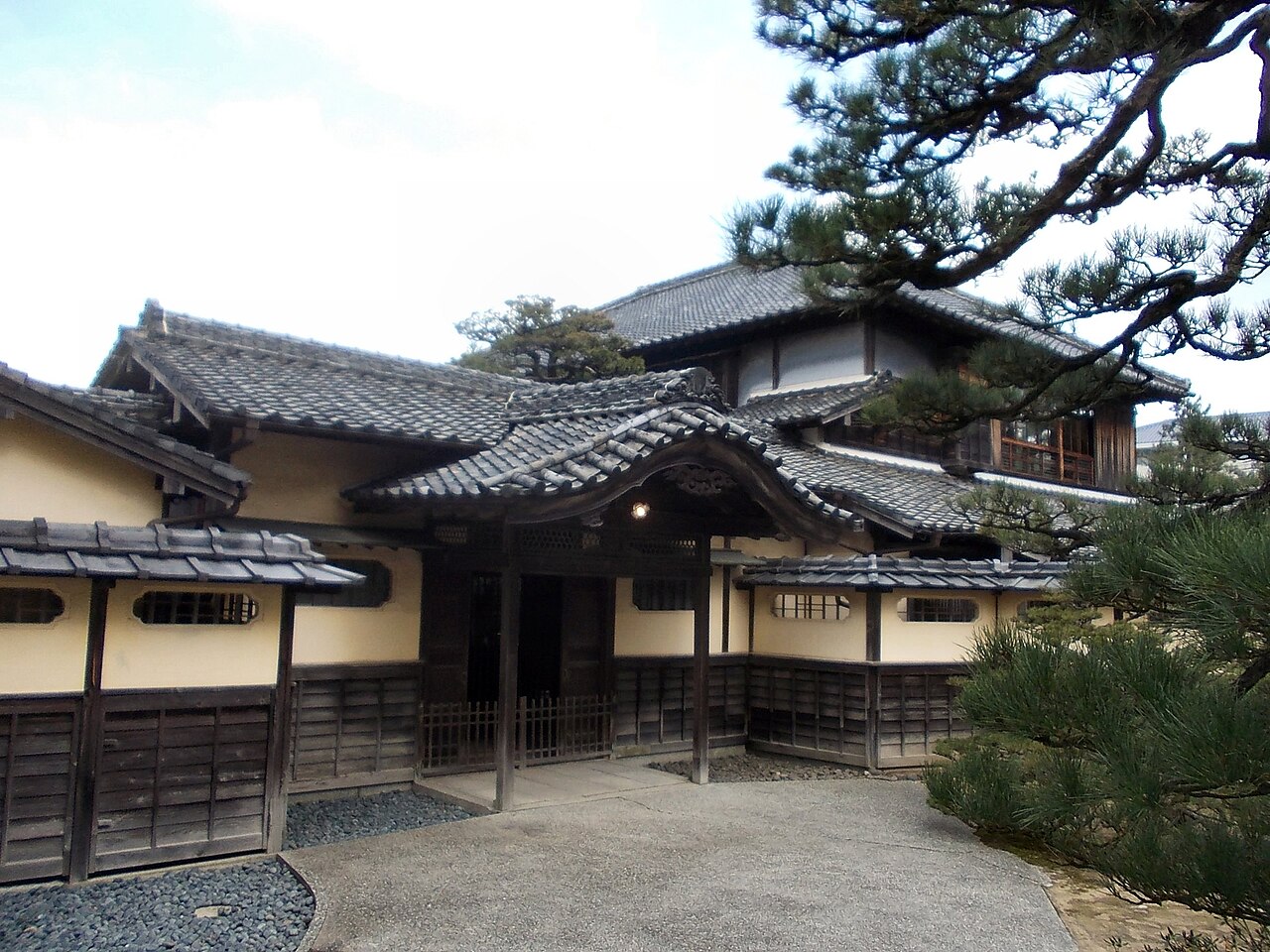 The tiled-roof gate of the Former Takatori Residence, with a curved gable over the entrance and pine branches reaching in from the right