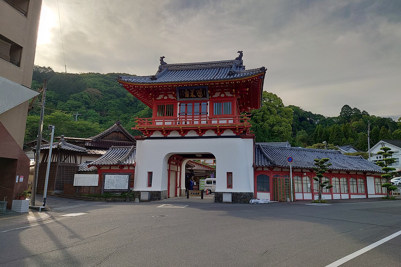 The two-storey vermilion gate of Takeo Onsen beneath a green hillside
