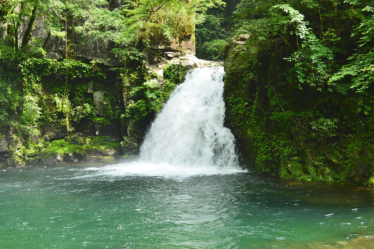 A wide white waterfall dropping over a rock ledge into a turquoise pool, framed by moss-covered rock and green leaves