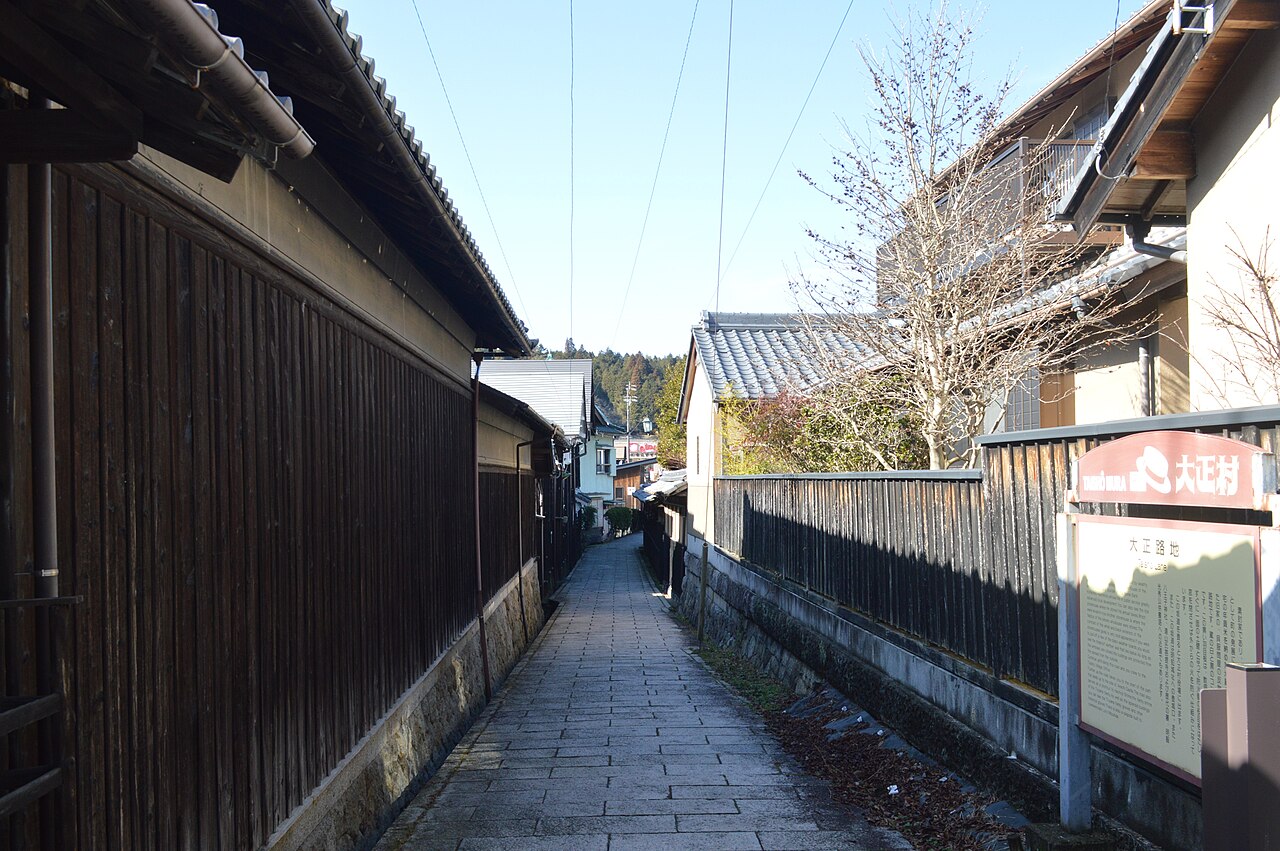 A narrow stone-paved lane between dark wooden walls in Akechi's Taisho Village, with a Taisho Mura sign board on the right under a clear sky
