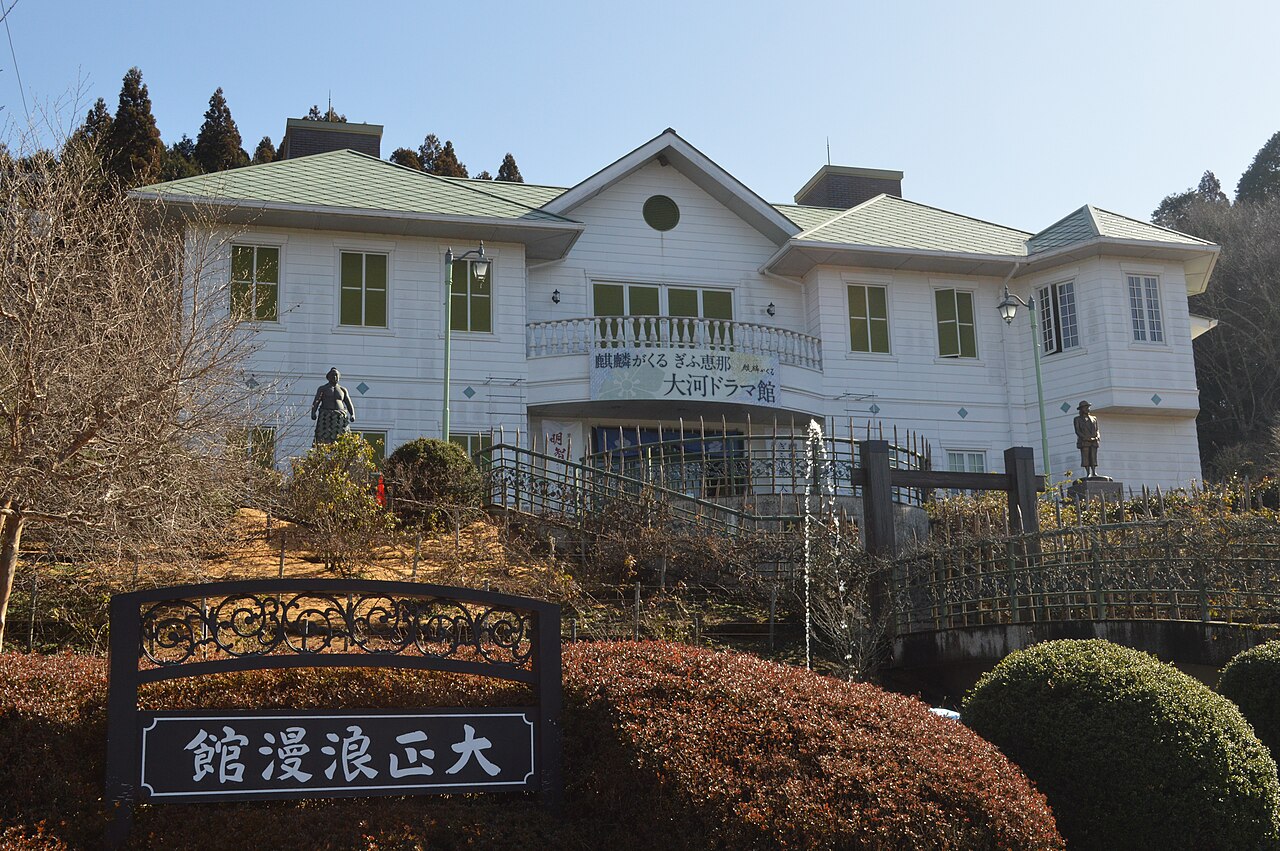 The white two-storey Taisho Roman Hall on a hillside above its name board, with bronze statues beside the entrance stairs