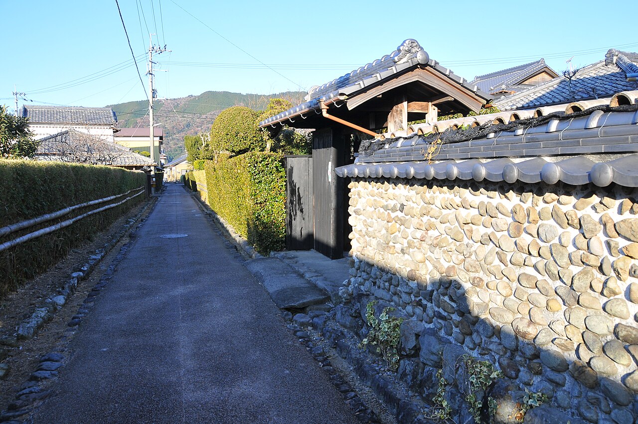 A quiet lane in the Doikachu samurai quarter of Aki, lined by trimmed hedges on one side and a wall of rounded river stones topped with tiles on the other