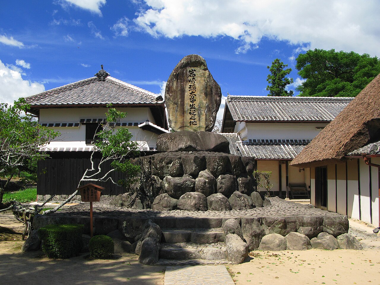 A large inscribed memorial stone on a stacked rock base in the courtyard of Iwasaki Yataro's birthplace in Aki, between a white-walled storehouse and a thatched-roof house
