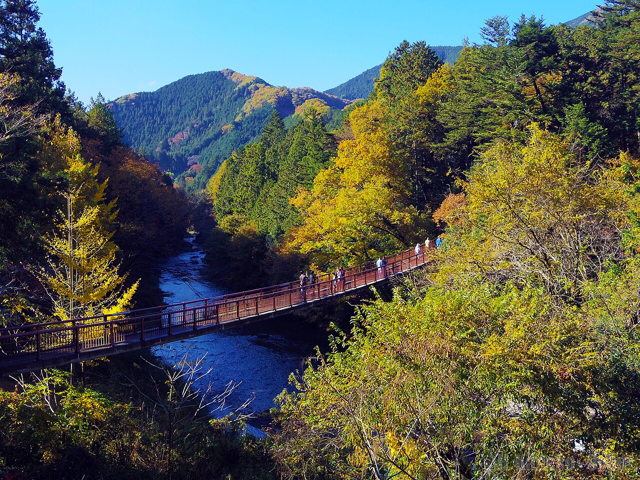 A red footbridge crossing the Akigawa river between wooded hills in autumn colour, with a few visitors pausing to look down at the clear water.