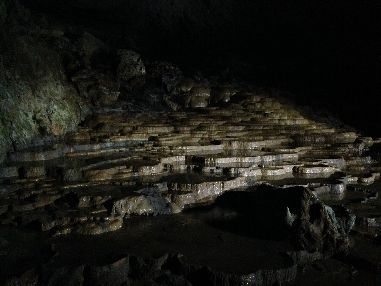 Terraced pools of pale limestone stacked like shallow dishes, lit by lamps inside the dark cave