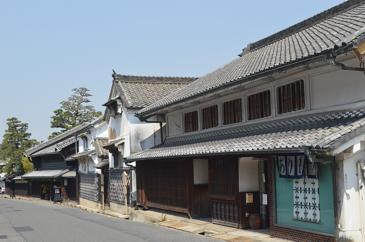 A row of old tile-roofed merchant houses with white plaster walls and latticed windows lining a quiet street in Arimatsu, under a clear blue sky, with a blue patterned noren at one doorway.