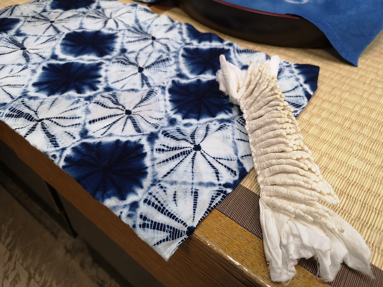 Blue and white tie-dyed cloth with round starburst patterns lying on a tatami edge, next to a tied bundle of cream-coloured cloth with rows of small knots.