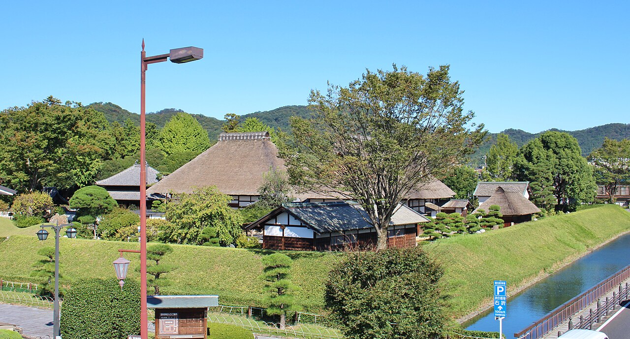 The grounds of Ashikaga Gakko, with a moat and bank in front of thatched-roof buildings and hills behind