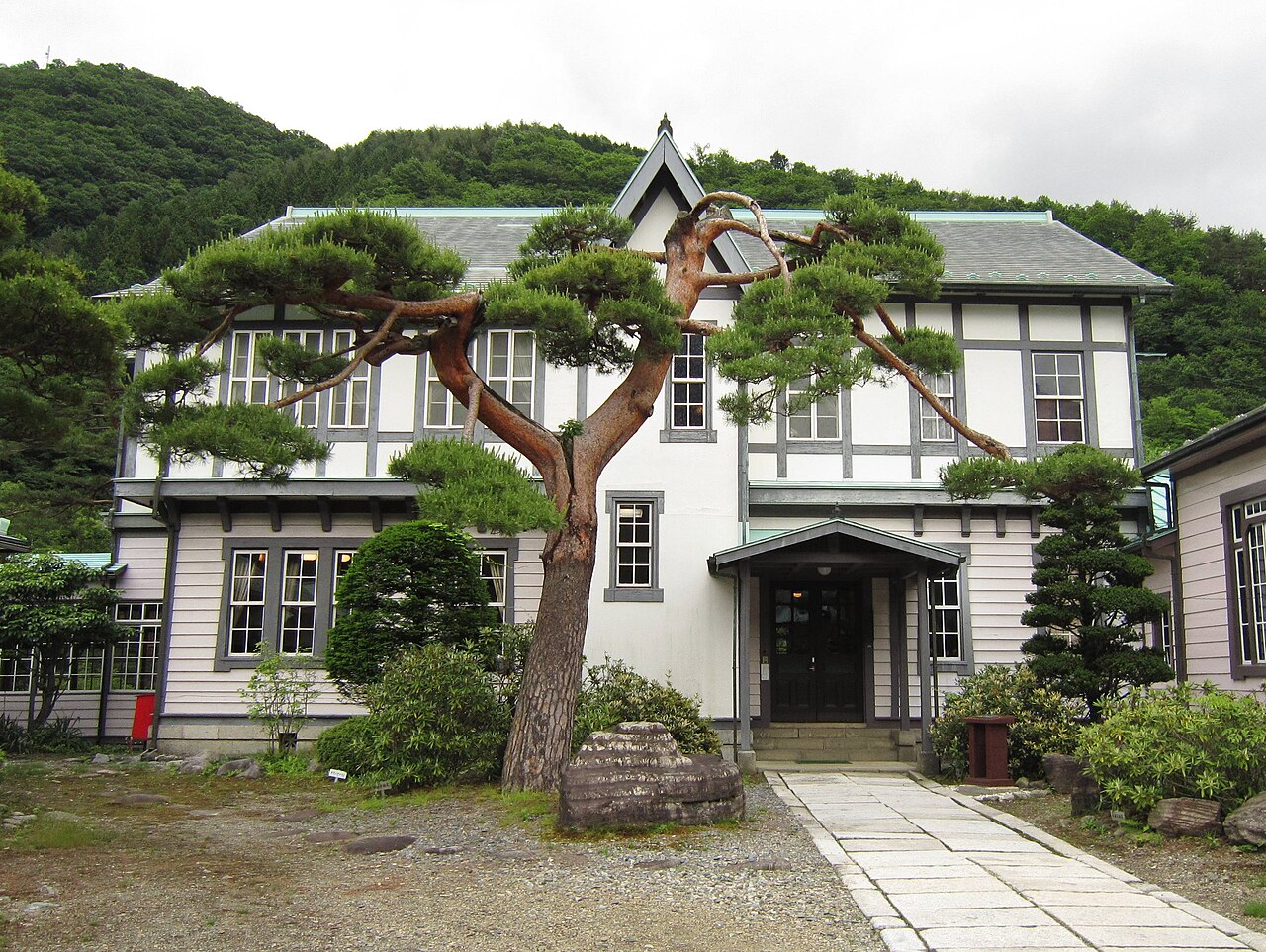 A two-storey white and grey Western-style timber house with a gabled porch stands behind a twisted pine tree and a stone path, with a forested hillside rising behind it