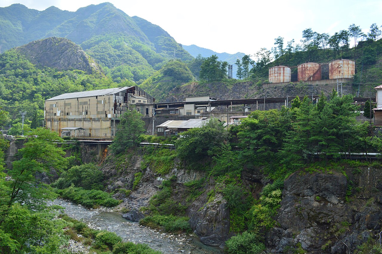 A large weathered factory hall and three rusty storage tanks sit on a ledge above a rocky stream in a sharply rising green mountain gorge