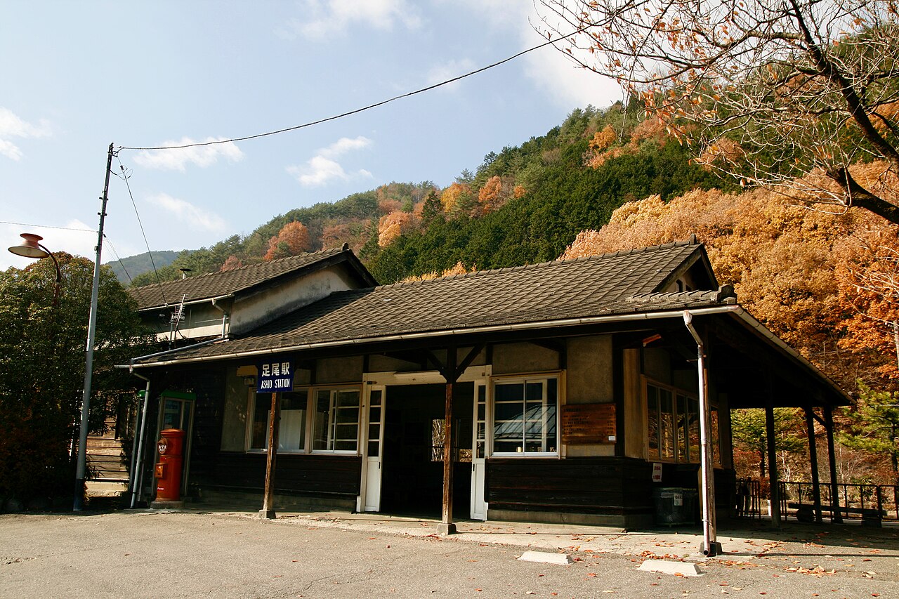 A low wooden station building with a dark tiled roof and a blue Ashio Station sign stands under hillsides of orange and green autumn leaves, with a small red postbox beside the entrance