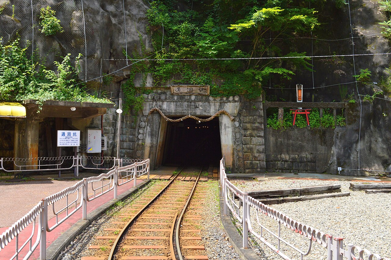 A dark mine tunnel mouth framed by stacked stone and a straw rope, with a narrow-gauge rail line running into it, a small red torii on the rock ledge above and green vines hanging down the rock face