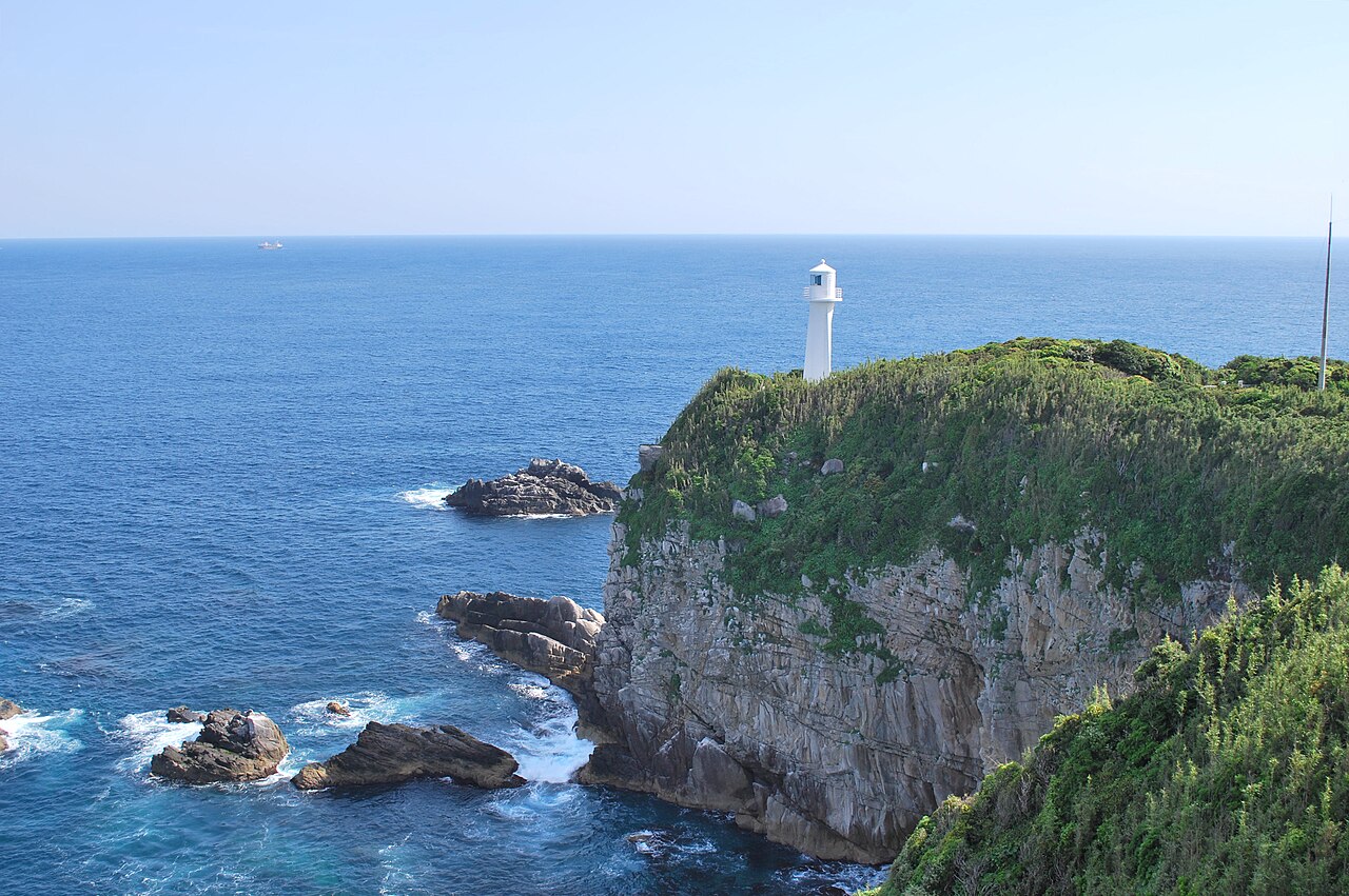 White Ashizuri Cape lighthouse standing on a green sea cliff above rocks and deep blue ocean