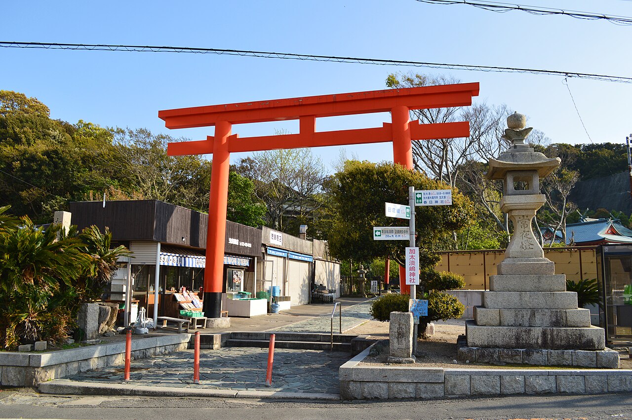 A vermilion torii gate and stone lantern at the approach to Awashima Shrine in Kada, with shops and trees beside the road.