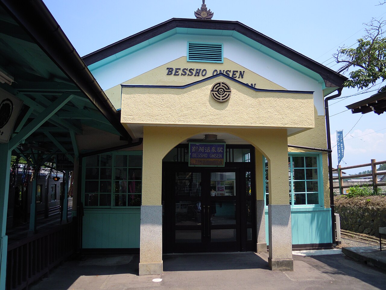The small pastel-yellow and turquoise station building of Bessho-Onsen Station with an arched entrance and the name in English letters on the gable, and a train waiting on the platform at left.